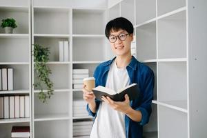 Masculin étudiant prise Remarques de une livre à bibliothèque, Jeune asiatique séance à bureau Faire affectations dans Université bibliothèque photo