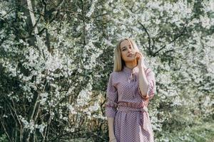 blond fille sur une printemps marcher dans le jardin avec Cerise fleurs. femelle portrait, fermer. une fille dans une rose polka point robe. photo