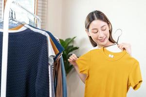 choix de vêtements, rien à porter. attrayant asiatique Jeune femme, fille essayer sur apparaître, content choisir robe, tenue sur cintre dans garde-robe dans pièce placard à maison. décider chemisier quoi à mettre sur lequel un photo