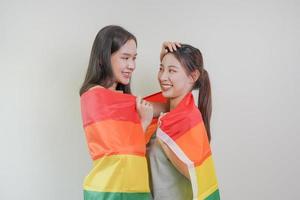 content lesbienne, magnifique asiatique Jeune deux femmes, fille gay, couple l'amour moment dépenses bien temps ensemble, en portant ou agitant lgbt arc-en-ciel, fierté drapeau sur isolé blanc mur Contexte ensemble à maison. photo