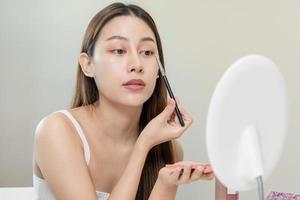 content beauté concept, mignonne asiatique Jeune femme, fille sourire, faire en haut visage par un le fard à paupières brosse sur sa yeux, à la recherche dans miroir dans Matin routine à maison. gens Regardez avec Naturel mode style. photo