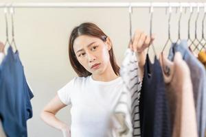 choix de vêtements, rien à porter. attrayant asiatique Jeune femme, fille essayer sur apparaître, content choisir robe, tenue sur cintre dans garde-robe dans pièce placard à maison. décider chemisier quoi à mettre sur lequel un photo