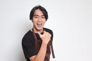 portrait de attrayant asiatique barista homme dans marron tablier avec pouce montrer du doigt une façon sur vide espace. La publicité concept. isolé image sur blanc Contexte photo