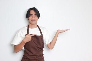 portrait de attrayant asiatique barista homme dans marron tablier montrant produit, montrer du doigt à quelque chose avec mains. La publicité concept. isolé image sur blanc Contexte photo