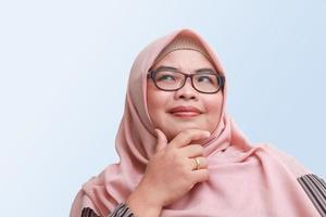 portrait de de bonne humeur asiatique femme avec hijab, permanent contre bleu arrière-plan, en pensant à propos question avec main sur menton photo