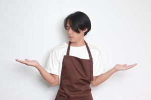portrait de attrayant asiatique barista homme dans marron tablier choisir entre 2 différent choix, en portant deux mains avec vide espace. décision et choix concept. isolé image sur blanc Contexte photo