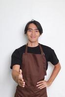 portrait de attrayant asiatique barista homme dans marron tablier atteindre en dehors pour une poignée de main et saluer quelqu'un. isolé image sur blanc Contexte photo