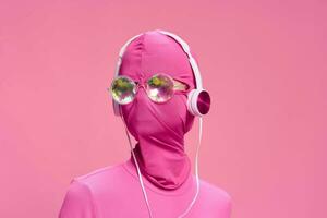 portrait de une homme portant une rose visage complet incognito masque avec écouteurs et brillant rond des lunettes de soleil sur une rose Contexte. conceptuel art la photographie total rose photo