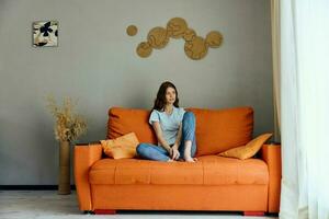 de bonne humeur femme sur le Orange canapé dans le du repos pièce posant inchangé photo