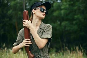 militaire femme portant foncé des lunettes détient une arme dans le sien mains Frais air vert salopette photo