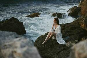 une femme est assis sur une falaise dans une blanc robe par le océan nuageux temps photo