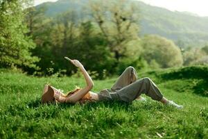 content femme blogueur mensonge sur le herbe dans le parc et souriant avec sa téléphone dans sa mains contre le toile de fond de une été Naturel paysage avec lumière du soleil photo