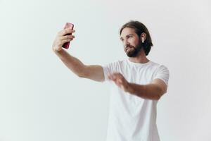 une Masculin blogueur enregistrement vidéo lui-même sur le sien téléphone et bavardage avec gens en ligne avec une sourire dans une blanc T-shirt contre une blanc mur photo