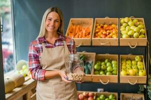 femme travaux dans des fruits et des légumes magasin. elle est en portant pot avec pistaches. photo