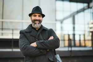 portrait de content homme d'affaire dans noir Vêtements permanent dans de face de entreprise bâtiment. noir barbu homme est portant hiver Vêtements et souriant. photo