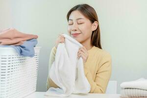 ressentir douceur, souriant asiatique Jeune femme, fille émouvant duveteux serviette coton, odeur Frais nettoyer vêtements sur table après la lessive, blanchisserie, sec. Ménage travail à maison. blanchisserie et femme de ménage. photo