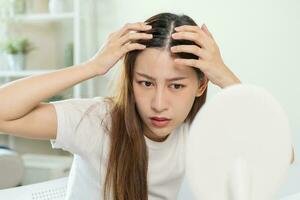 endommagé cheveux, visage sérieux asiatique Jeune femme inquiéter à la recherche à cuir chevelu dans miroir, main dans Pause dans de face cheveux perte, mince problème symptôme à maison. santé se soucier shampooing beauté, isolé sur Contexte. photo