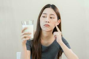 allergie asiatique Jeune femme, fille en regardant, en portant verre de lait, visage dans en pensant avant boisson Lait comme il mai dérangé sa estomac mal, douleur. lactose intolérance et laitier nourriture , santé problème concept. photo