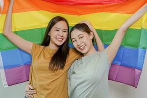 content lesbienne, magnifique asiatique Jeune deux femmes, fille gay, couple l'amour moment dépenses bien temps ensemble, en portant ou agitant lgbt arc-en-ciel, fierté drapeau sur isolé blanc mur Contexte ensemble à maison. photo