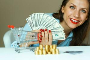 Jeune souriant femme en portant espèces, et sur le table sont pièces de monnaie, crédit carte et le achats Chariot photo