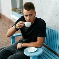 Beau homme avec le coiffure dans une noir chemise est assis sur une bleu banc et boisson café. photo
