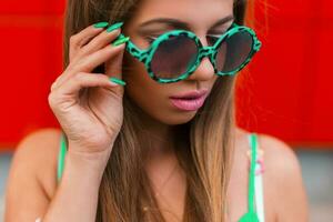 fermer élégant portrait de une jolie femme avec rose lèvres et vert rond des lunettes de soleil sur une rouge Contexte. photo