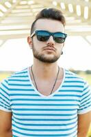 Beau homme dans une T-shirt avec le barbe dans des lunettes de soleil permanent sur le plage. photo