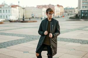 élégant gars avec des lunettes dans branché veste et déchiré jeans avec noir baskets dans le ville photo