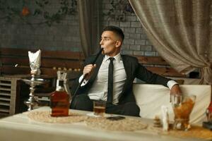 Jeune élégant homme dans une costume repose dans le bar. homme en buvant whisky et fumeur une narguilé. photo