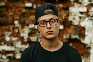 Beau gars avec des lunettes dans une noir base-ball casquette et noir T-shirt permanent sur une brique mur Contexte photo