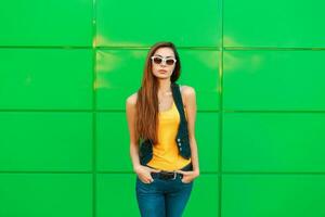 Jeune à la mode femme dans une Jaune T-shirt et denim gilet posant près le vert mur. photo