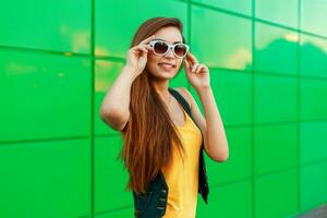 content de bonne humeur élégant femme dans des lunettes de soleil posant près le brillant vert mur. photo