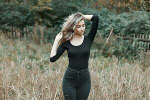 magnifique fille avec cheveux dans une noir chemise et noir jeans permanent dans une champ avec herbe. photo