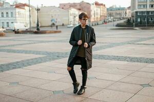 élégant gars avec des lunettes dans branché veste et déchiré jeans avec noir baskets dans le ville photo