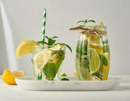 limonade dans une transparent verre avec citron, chaux, Romarin brins et menthe feuilles sur une blanc Contexte photo
