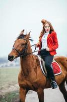 Fille jockey aux cheveux roux dans un cardigan rouge et des bottes noires avec un cheval photo