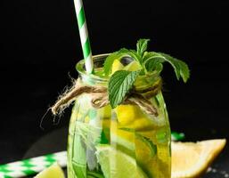 limonade dans une transparent verre avec citron, chaux, Romarin brins et menthe feuilles sur une noir Contexte photo