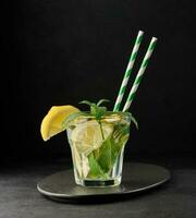 limonade dans une transparent verre avec citron, chaux, Romarin brins et menthe feuilles sur une noir Contexte photo