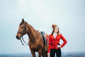 Fille jockey aux cheveux roux dans un cardigan rouge et des bottes noires avec un cheval photo