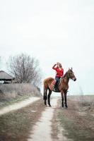 Fille jockey aux cheveux roux dans un cardigan rouge et des bottes noires avec un cheval photo