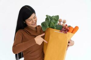 content asiatique femme souriant et point le doigt à une papier sac et porte une achats sac après le courrier de épicerie venu à livrer à maison. concept de supermarché livraison pour une Nouveau mode de vie. photo