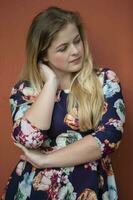 une pensif blond fille avec bleu yeux dans une bleu robe avec une floral impression des stands près une Bourgogne mur. photo