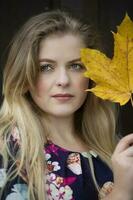 proche en haut est le visage de une magnifique fille en portant une Jaune érable feuille. aux yeux bleus blond dans l'automne. photo