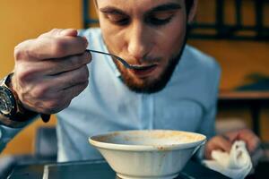 une Beau homme avec une barbe mange Bortsch dans une assiette à une table dans une café et une regarder sur le sien main photo