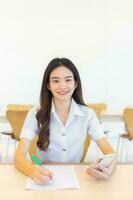 Jeune asiatique femme étudiant dans uniforme en utilisant téléphone intelligent et l'écriture quelque chose à propos y travailler sont beaucoup les documents sur le table sa visage avec souriant dans travail à à chercher information pour étude photo