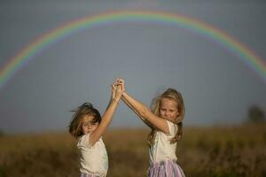 deux peu les filles supporter dans une été champ avec leur mains en haut en dessous de une arc-en-ciel. sœurs. photo