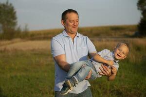 content papa pièces avec le sien fils sur le Contexte de le champ. le joyeux grand-père virevolte le sien petit fils dans le sien bras. photo