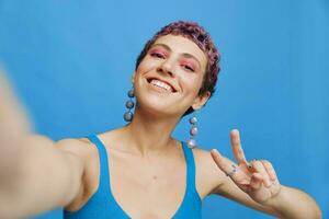 Jeune des sports femme mode blogueur prend une image de se sur le téléphone dans bleu tenue de sport souriant et à la recherche à le caméra sur une bleu monochrome Contexte photo