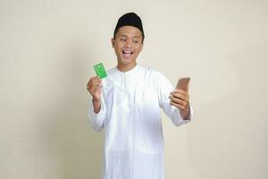 portrait de attrayant asiatique musulman homme dans blanc chemise avec calotte en portant une mobile téléphone et en présentant crédit carte. isolé image sur gris Contexte photo