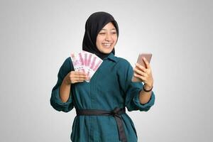 portrait de excité asiatique hijab femme dans décontractée tenue montrant un cent mille Rupiah tandis que en portant une mobile téléphone. financier et des économies concept. isolé image sur blanc Contexte photo